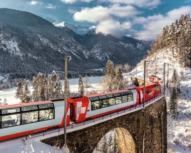 A red train is crossing a bridge through a snowy mountain landscape.