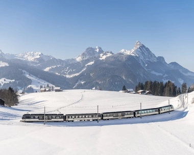 A winter mountain landscape with a train traveling through snowy fields.