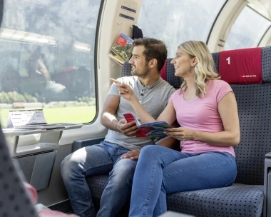 A couple sits on a train, looking out the window and holding travel guides in their hands.