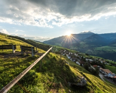 Eine Bank auf einem Hügel mit Blick auf ein Dorf im Tal, Sonnenuntergang hinter Bergen.