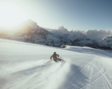 A skier on a freshly groomed slope with snow-covered mountains in the background.