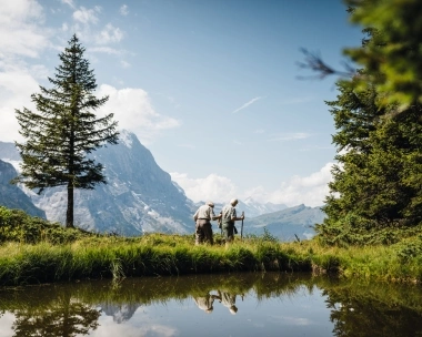 Deux personnes randonnent sur un sentier de montagne avec des arbres et un étang au premier plan.