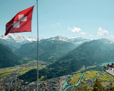 A group of people on a viewing platform overlooking a valley and mountains, next to a Swiss flag.