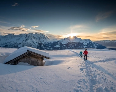 Two people hiking in the snow at sunset, mountains in the background.