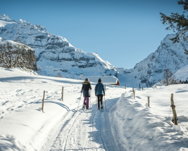 Two people are hiking on a snowy path in a winter landscape with mountains in the background.