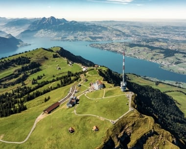 Vue aérienne d'un paysage montagneux vert avec une grande tour de transmission et un lac en arrière-plan.