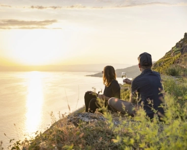 Zwei Personen sitzen bei Sonnenuntergang mit einem Glas Wein am Hang und blicken aufs Meer.