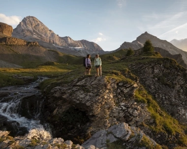 Due persone in piedi su una scogliera rocciosa in un paesaggio montuoso al tramonto.