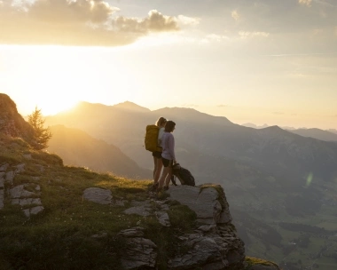 Zwei Wanderer stehen auf einem Berggipfel bei Sonnenuntergang.