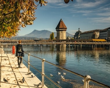 Une femme se promène le long de la rive, avec le Pont de la Chapelle et la Tour de l'eau à Lucerne en arrière-plan; montagnes en arrière-plan.