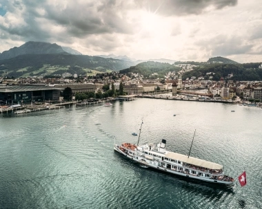 Eine Stadt am See mit Bergen im Hintergrund, ein Schiff fährt über das Wasser.