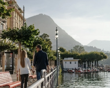 A woman and a man walk along a waterfront promenade, with trees and a mountain visible in the background.