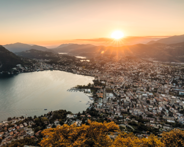 View of a city by a lake with mountains in the background at sunset.