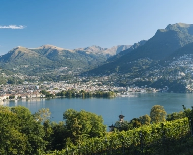 A city lies next to a large, mountain-surrounded lake under a blue sky.