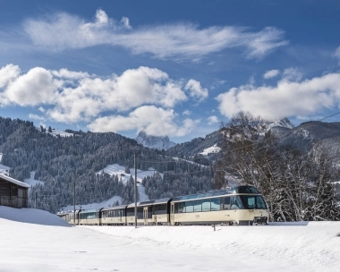 Eine Bahn fährt durch eine verschneite Berglandschaft unter einem blauen Himmel.