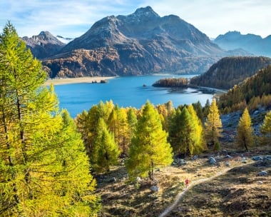 A picturesque mountain landscape with a lake, surrounded by autumn-colored trees and a hiker on a trail.