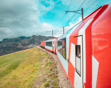 Ein roter Zug fährt auf einer Gebirgsstrecke, umgeben von grünen Wiesen und dramatischen Wolken.