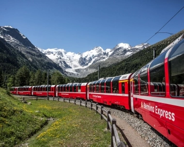 A red Bernina Express train travels through an alpine landscape with snow-covered mountains in the background.