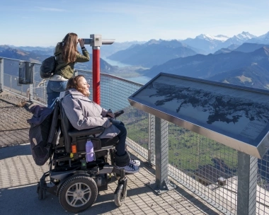 A woman in a wheelchair enjoys the mountain view while another person looks through binoculars.