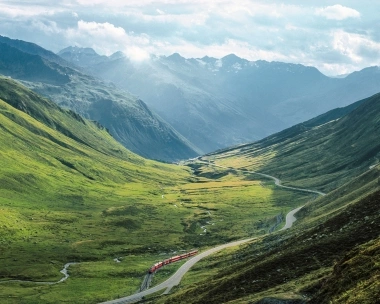 A red train travels through a green Alpine valley with a winding road and mountain views in the background.