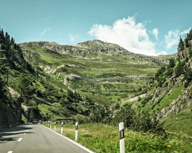 Eine kurvige Bergstrasse, umgeben von grünen Hügeln und Felsen unter blauem Himmel.