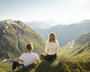 Due persone sono sedute su un prato con vista su un paesaggio montuoso.