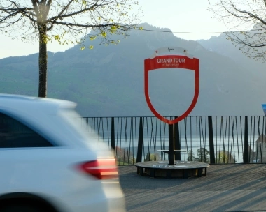 Eine weisse Limousine fährt an einem 'Grand Tour of Switzerland' Schild vorbei, im Hintergrund Berge und ein See.