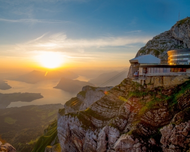 Sonnenuntergang über einer Gebirgslandschaft mit einem Gebäude am Berghang.