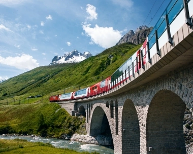 Un train rouge traverse un viaduc à travers un paysage montagneux vert avec des sommets enneigés.