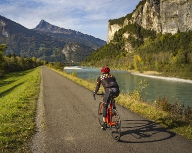 Ein Radfahrer auf einem Weg entlang eines Flusses mit Bergen im Hintergrund.