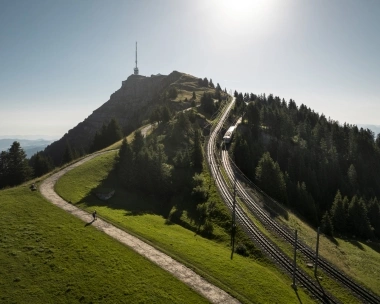 Eine Berglandschaft mit einer Eisenbahnstrecke, die zu einem Gipfel mit einem Sendemast führt, umgeben von Bäumen und Wiesen.