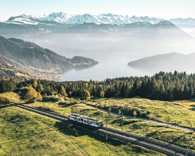 A train travels over green hills with mountains and a lake in the background.