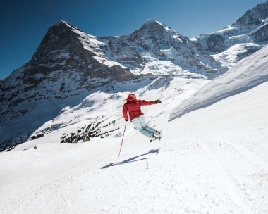 Ein Skifahrer in roter Jacke springt auf einer verschneiten Piste vor einer beeindruckenden Bergkulisse.