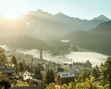 Tramonto su una cittadina alpina con chiesa, circondata da montagne e un lago.