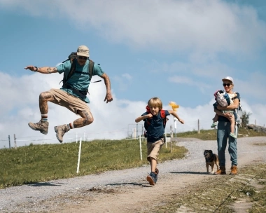 Une famille marche sur un chemin, tandis qu'un homme et un enfant sautent joyeusement ; une femme porte un petit enfant.