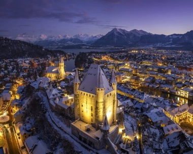 A winter city at night, illuminated buildings, snow-covered roofs, and mountains in the background.