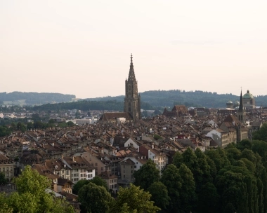 A city view at sunset with a tall church tower and surrounding green landscape.