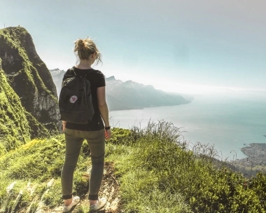 Una donna con lo zaino in piedi su una montagna guarda un lago e il paesaggio circostante.