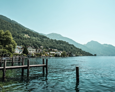 A tranquil lake landscape with houses on the shore and wooded mountains in the background.