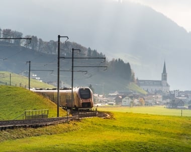 Zug auf einer Eisenbahnstrecke durch eine grüne Landschaft mit einem Dorf im Hintergrund.