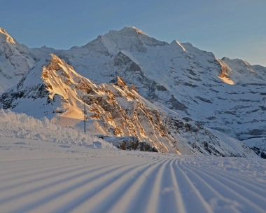 Sunlit, snowy mountain landscape with a groomed ski slope in the foreground.