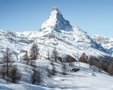 Eine verschneite Berglandschaft mit einem markanten Gipfel im Hintergrund und einem leeren Sessellift im Vordergrund.