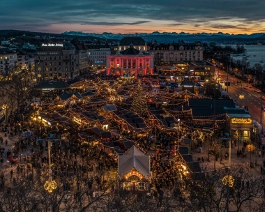 Aerial view of a festively lit Christmas market at dusk, surrounded by buildings with a view of mountains and a lake.