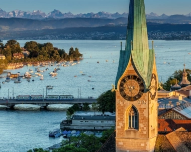 A church with a clock tower, view of the lake with boats and mountains in the background.