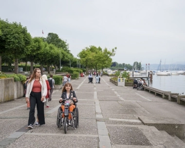 Eine Frau geht neben einer Person, die ein Handbike benutzt, entlang einer Promenade am Wasser; im Hintergrund sind Boote zu sehen.