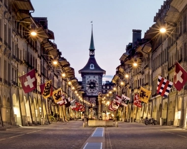 An illuminated street in Bern with the Zytglogge Tower and Swiss flags.