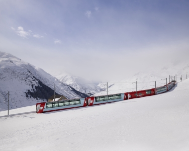 Un train rouge traverse des montagnes enneigées sous un ciel bleu.