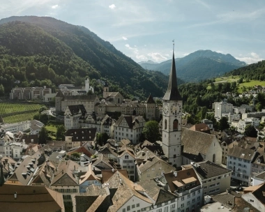 Eine Stadtansicht mit einer Kirche im Vordergrund, umgeben von historischen Gebäuden und grünen Hügeln im Hintergrund.