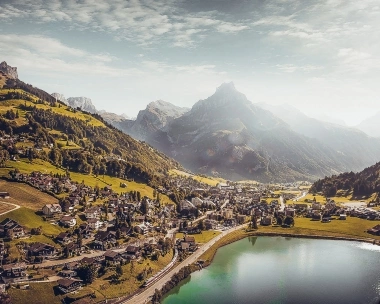 Villaggio con case vicino a un lago, circondato da colline verdi e alte montagne sullo sfondo.