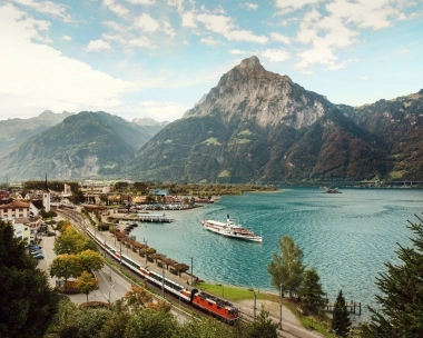 Mountain landscape with lake, train, and ship in the foreground.